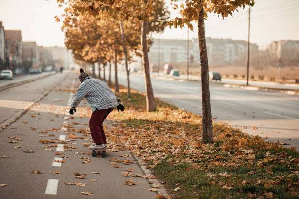Person in beanie riding a longboard on a leaf-strewn autumn path, trees line the road.
