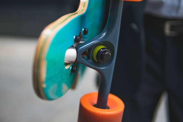 Close-up of a longboard's teal deck, grey truck, and vibrant orange wheel.