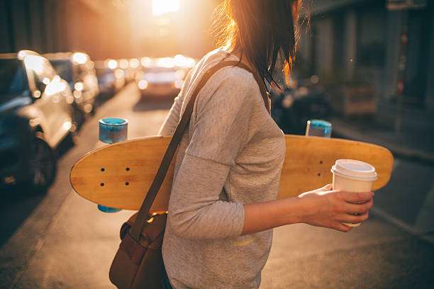 Woman holding a wooden longboard and coffee cup, walking on a sunny city street.