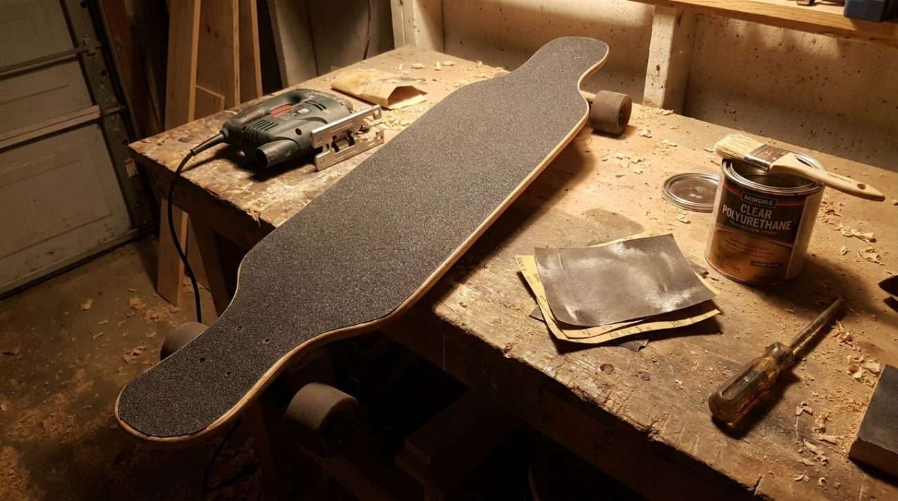 Finished DIY longboard deck with grip tape applied, leaning against a wall beside the tools used to build it